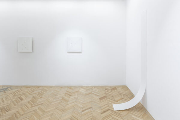 Róg pomieszczenia z białymi ścianami i drewnianą podłogą. Na jednej ze ścian wisi biała pleksi zawijająca się przy podłodze, na drugiej dwa białe obrazy w równych odległościach.