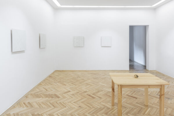 Drewniany stół z pożółkłym papierem, zgniecionym w kulkę, w pomieszczeniu z drewnianą podłogą i białymi ścianami, na których wiszą białe obrazy w równych odstępach oraz wejściem do sali w prawej części ściany.