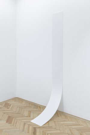 Róg pomieszczenia z białymi ścianami i drewnianą podłogą. Na jednej ze ścian wisi biała pleksi zawijająca się przy podłodze.