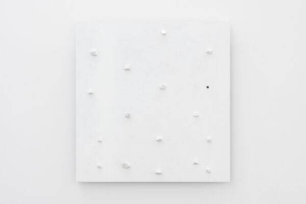 Biały, kwadratowy obraz z białymi kamykami umieszonymi w nierównych odstępach na całej jego powierzchni i jedną, czarną dziurką. Wisi na białej ścianie.