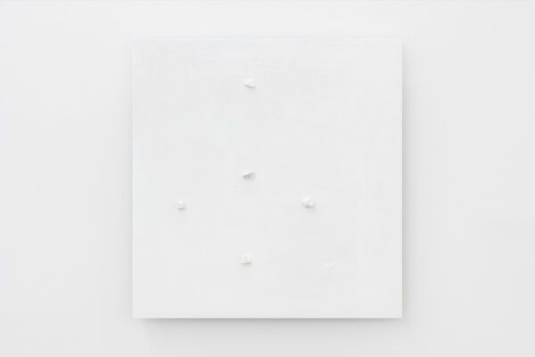 Biały, kwadratowy obraz z pięcioma białymi kamykami umieszonymi w nierównych odległościach, przypominając krzyż. Wisi na białej ścianie.