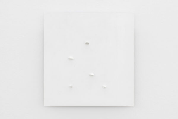 Biały, kwadratowy obraz z pięcioma białymi kamykami umieszonymi w nierównych odległościach w okolicach środka pracy. Wisi na białej ścianie.