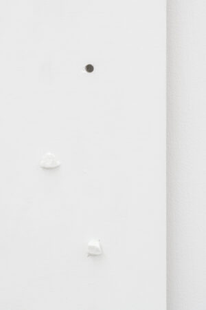 Fragment krawędzi białego obrazu z dwoma białymi kamykami i małą, czarną dziurką, na tle białej ściany.
