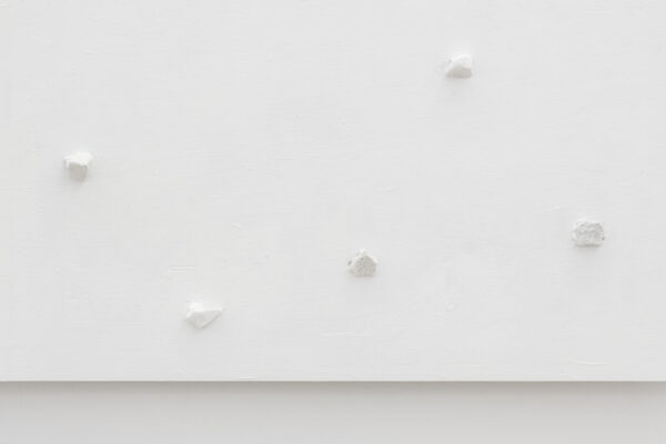 Fragment krawędzi białego obrazu z pięcioma białymi kamykami rozmieszczonymi w nierównych odległościach, na tle białej ściany.
