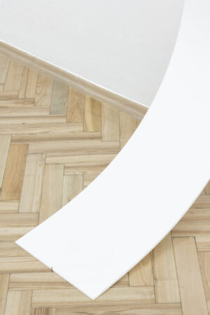 Biała pleksi schodząca pod łagodnym kątem z białej ściany do drewnianej podłogi.