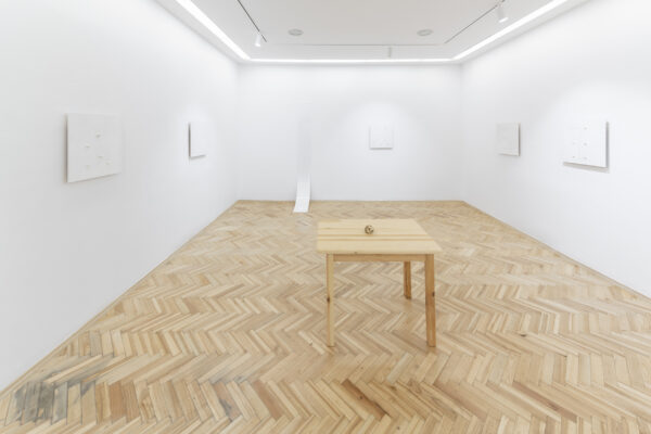 Drewniany stół ze zgniecionym papierem w kształcie kulki w pomieszczeniu z drewnianą podłogą i białymi ścianami, na których wiszą białe obrazy w równych odstępach oraz jedna podłużna pleksi w białym kolorze zawijająca się przy podłodze.